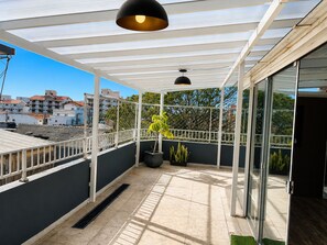 Ein überdachter Balkon mit einem Blumentopf, einer schwarzen Pendelleuchte und Blick auf Gebäude im Hintergrund.