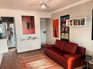 A living room with a red sofa, a ceiling fan, and a bookshelf. - Penthouse with ocean view, one block from the beach, with private garage. (Florianópolis)