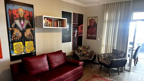 A living room with a red sofa, a patterned armchair, and a bookshelf with various books and posters. - Penthouse with ocean view, one block from the beach, with private garage. (Florianópolis)