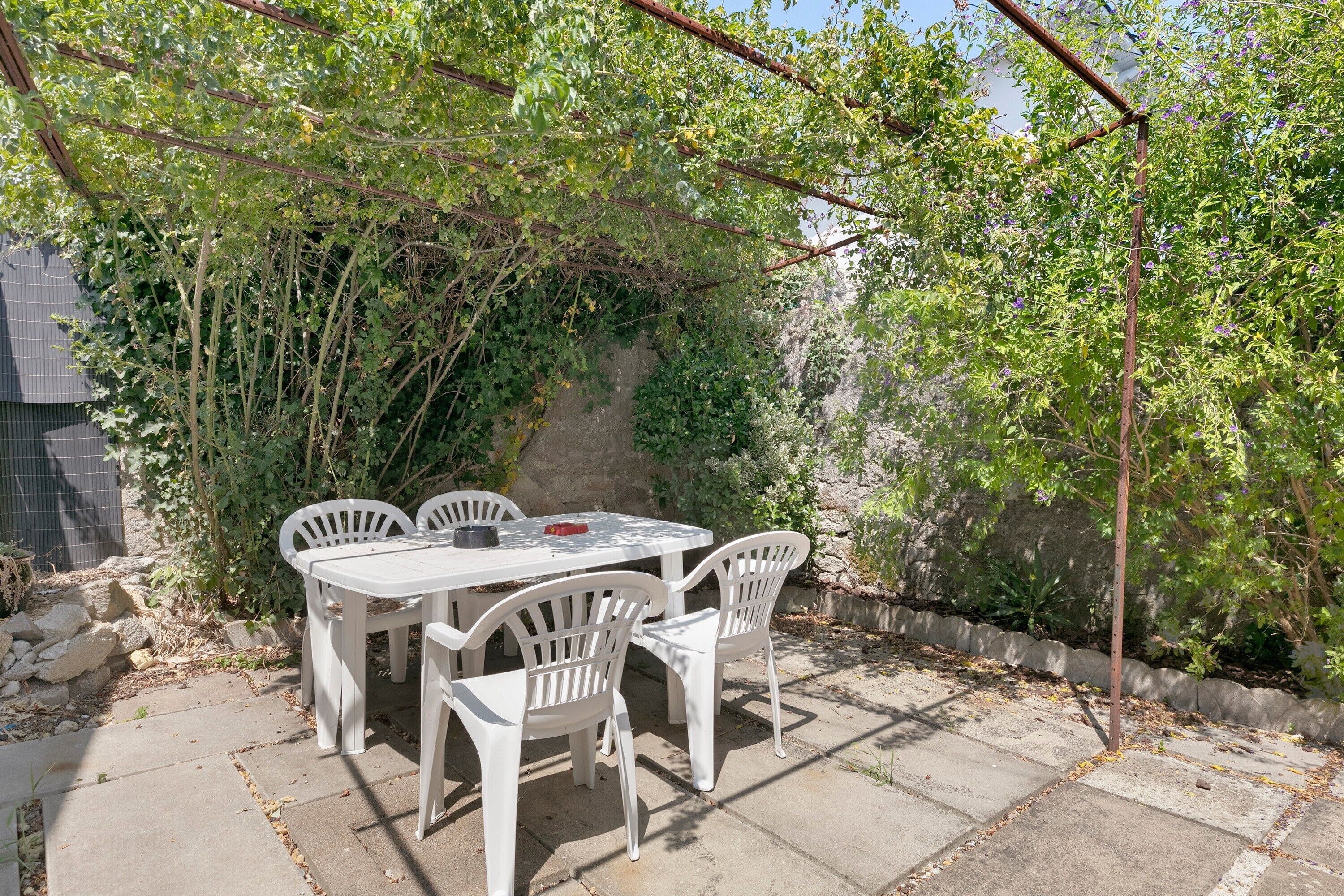 An outdoor patio with a table and chairs, surrounded by greenery and a stone wall.