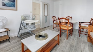 A room with a dining table, chairs, a baby stroller, a fan, and a door leading to another room. - Hoedic – Apartment for 6 Guests with Wi-Fi (Le Croisic)