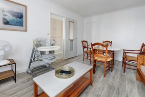 A room with a dining table, chairs, a baby stroller, a fan, and a door leading to another room. - Hoedic – Apartment for 6 Guests with Wi-Fi (Le Croisic)