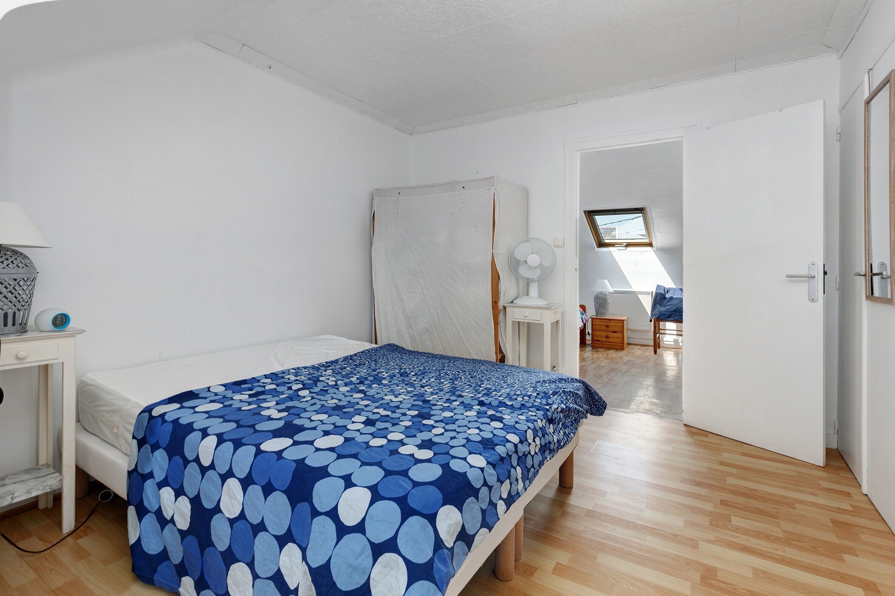 Quarto com cama, mesa de cabeceira, abajur, janela e porta que dá para outro cômodo.