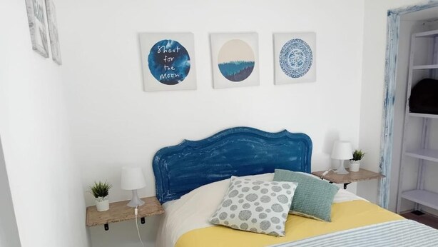 Um quarto com cabeceira azul, cama com colcha amarela e listrada, duas mesas de cabeceira com abajures e pequenas plantas, e três quadros na parede.