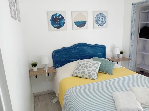 A bedroom with a blue headboard, a bed with a yellow and striped bedspread, two bedside tables with lamps and small plants, and three wall art pieces.