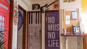 Quarto com uma porta, um letreiro 'NO MUS NO LIFE', um objeto decorativo de prancha de surf, uma guitarra e um painel de cortiça com anotações e porta-retrato.