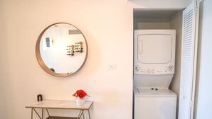 A compact room with a round mirror, a small table with a vase, and a built-in washer and dryer. - 1BR  Intracoastal View - Resort & Pool (Hallandale Beach)