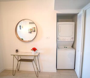 A compact room with a round mirror, a small table with a vase, and a built-in washer and dryer. - 1BR  Intracoastal View - Resort & Pool (Hallandale Beach)