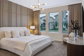 Ein Hotelzimmer mit einem großen Bett, einem Nachttisch, einer Lampe, einem Sessel, einem runden Tisch und Blick durch die Fenster hinaus.