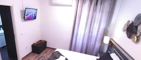 A bedroom with a bed, a flat-screen TV mounted on the wall, a small wooden stool, and a curtain.