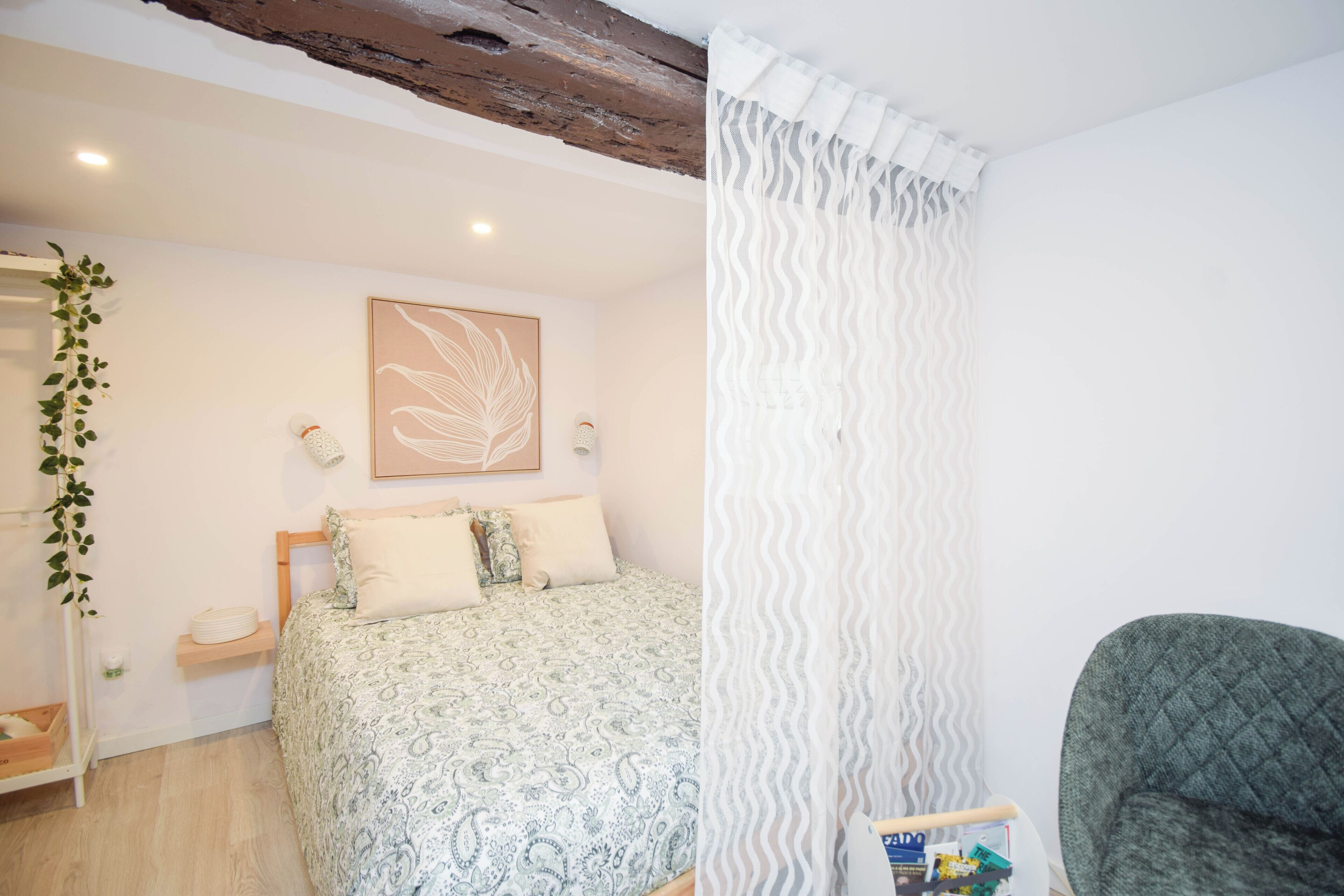 A bedroom with a bed, a bedside table, a chair, a wall-mounted shelf, and a hanging plant.
