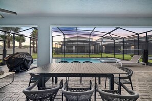 Une terrasse extérieure avec une piscine, un espace salon abrité et une vue sur une maison, le tout visible à travers une zone protégée par un moustiquaire.
