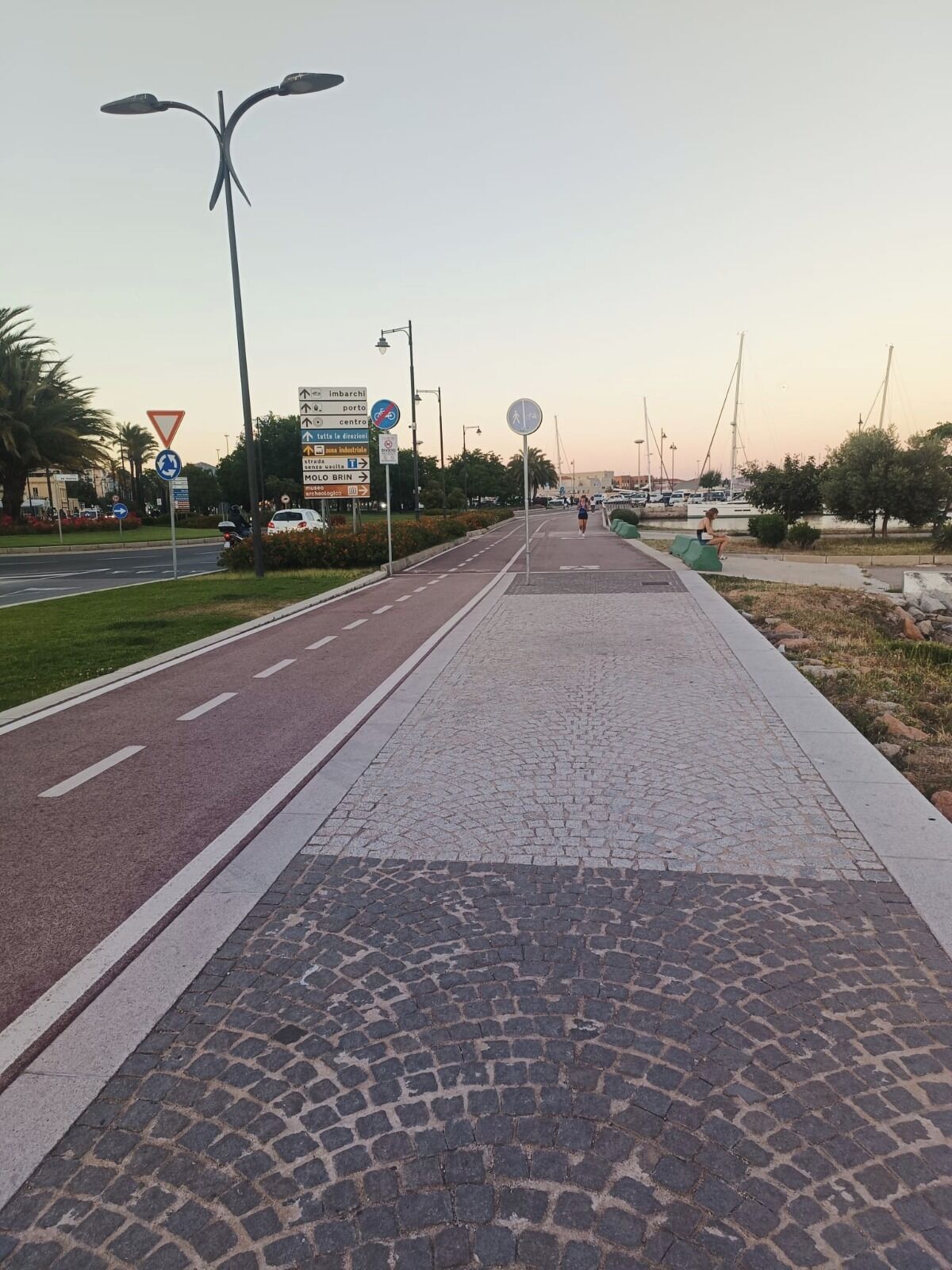 A paved walkway with a cobblestone section, a road with a bicycle lane, and directional signs.