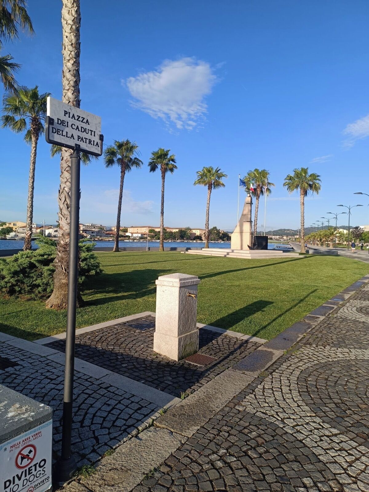 A paved area with a sign reading 'Piazza dei Caduti della Patria,' palm trees, a statue, and a body of water in the background.