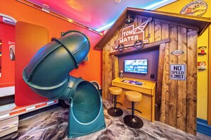 Una habitación con un tobogán grande, una pared de madera con un letrero de 'TOWING & SALVAGE', un cartel de 'NO SPEED LIMIT' y una zona de bar con taburetes.