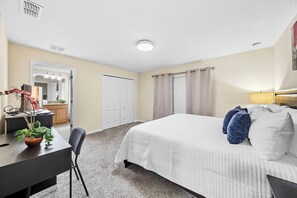 Une chambre d’hôtel comprenant un lit, un bureau, une chaise, une plante, un miroir, une armoire et une fenêtre avec des rideaux.