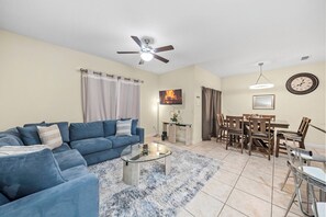A living room with a blue sofa, a glass coffee table, a dining area with wooden chairs, a ceiling fan, and a clock on the wall. - 4BR house with private pool in Paradise Palms (Kissimmee)