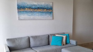 Een grijze modulaire bank met een passende voetenbankje, een blauw-wit abstract schilderij aan de muur, en een ronde houten salontafel.