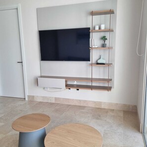 A modern living room with a flat-screen TV mounted on the wall, wooden shelves, a small plant, and two round tables. - Buenaventura Luxury Condo Beach Club • Golf (Río Hato)