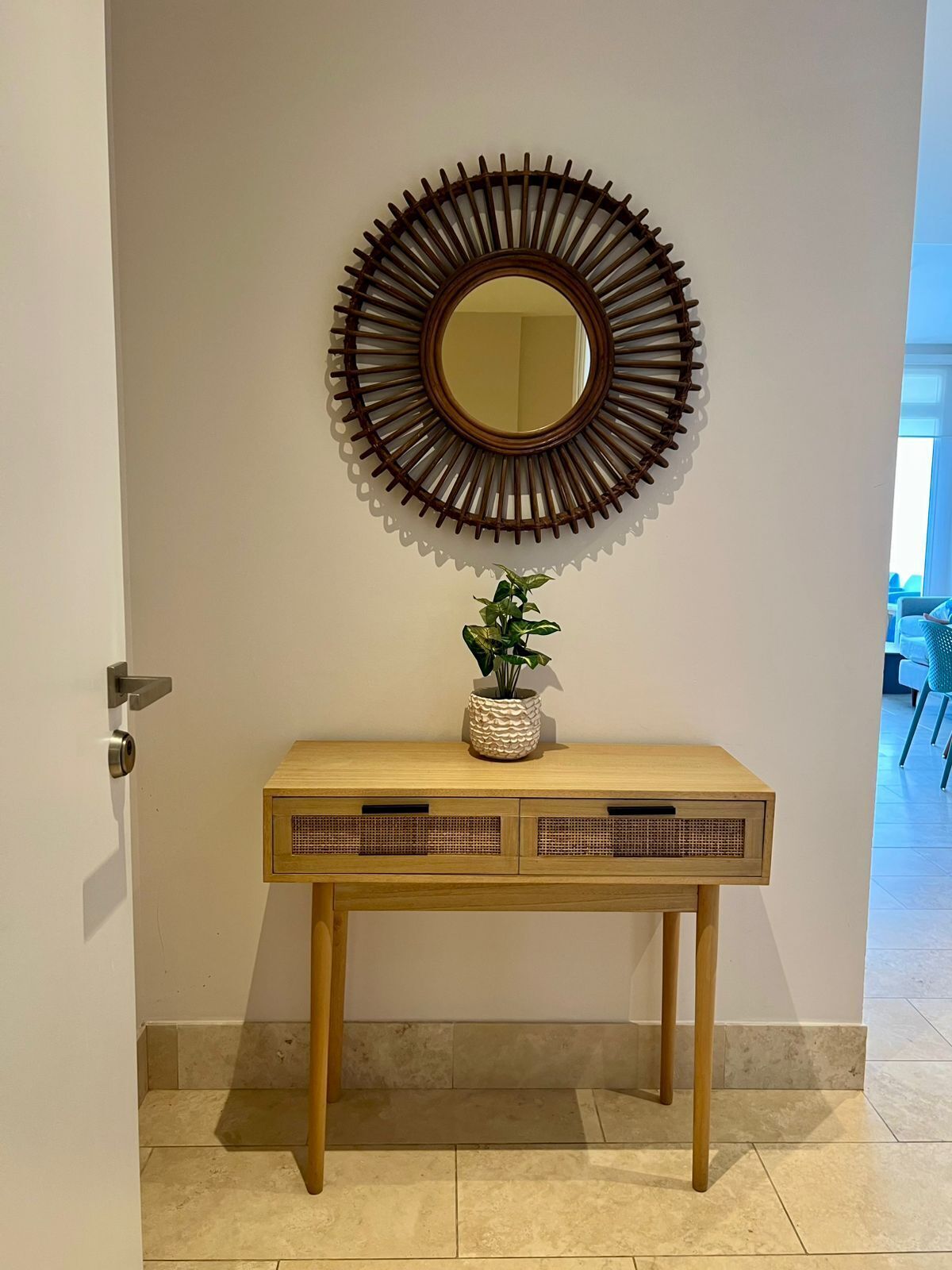 Une console en bois, surmontée d’un miroir décoratif, avec une plante en pot posée sur le plateau et une porte située à gauche.