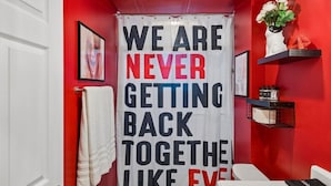 A bathroom with a red wall, a white shower curtain with bold text, a white towel, and a shelf with items. - Music Row | 5 Legend-Themed Suites | Sleeps 26 (Nashville)