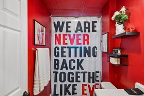 A bathroom with a red wall, a white shower curtain with bold text, a white towel, and a shelf with items. - Music Row | 5 Legend-Themed Suites | Sleeps 26 (Nashville)