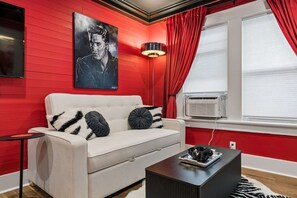 Ein Zimmer mit weißem Sofa, roten Wänden, einem schwarz-weiß gerahmten Porträt, einer Stehlampe und einem Fenster mit roten Vorhängen.