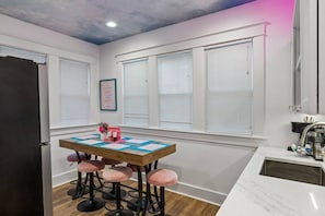 A kitchen with a wooden table, four bar stools, and a window with blinds. - Music Row | 5 Legend-Themed Suites | Sleeps 26 (Nashville)