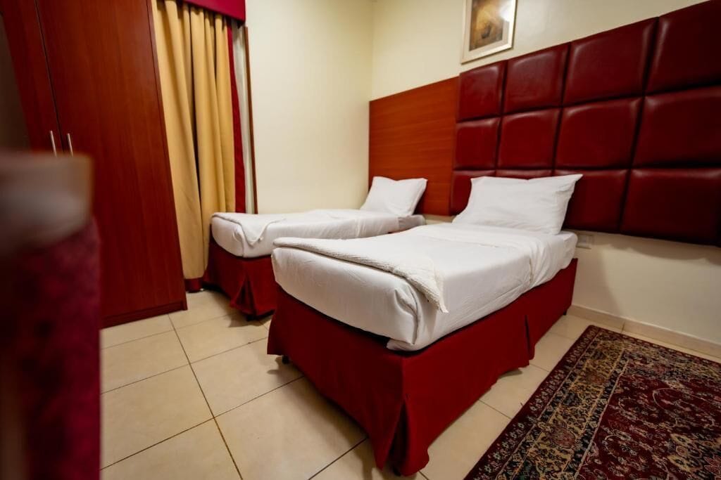 A hotel room with two beds, a red headboard, a patterned rug, and a door with curtains.