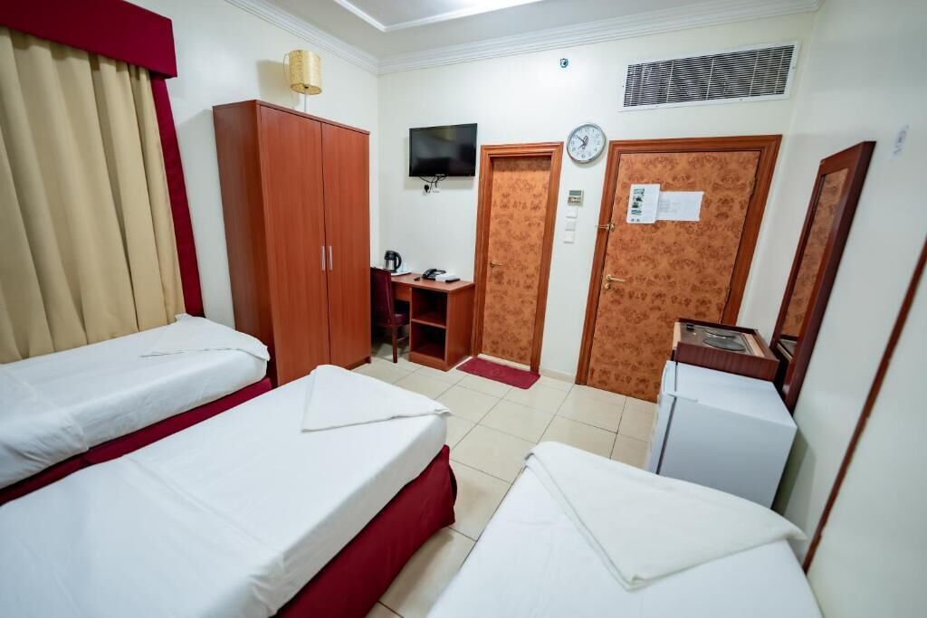 A hotel room with two single beds, a wooden wardrobe, a desk with a chair, a television, a clock, and a small refrigerator.