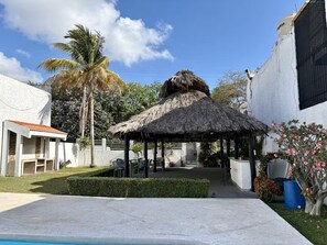 A pool area with a thatched-roof gazebo, palm trees, and a white building with a red-tiled roof. - Casa Céntrica, Cerca de la Playa. Acogedora con alberca en la residencial. (Cancún, QROO)