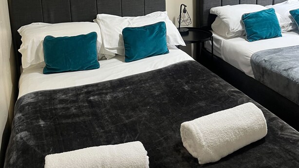 A hotel room with two beds, each with a dark headboard, white bedding, and teal pillows. There is a wooden railing above the beds, and a small table with a lamp. - Tudors eSuites - Budget Apartments (Birmingham)