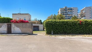 Ein Wohngebiet mit befestigter Zuwegung, einer Hecke mit roten Blüten und mehrstöckigen Gebäuden im Hintergrund.