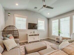 Un salon comprenant un canapé, un pouf, un téléviseur fixé au mur, un ventilateur de plafond et de grandes fenêtres équipées de stores.
