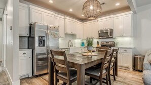 Una cocina moderna con armarios blancos, electrodomésticos de acero inoxidable, una mesa de comedor de madera y una lámpara colgante.