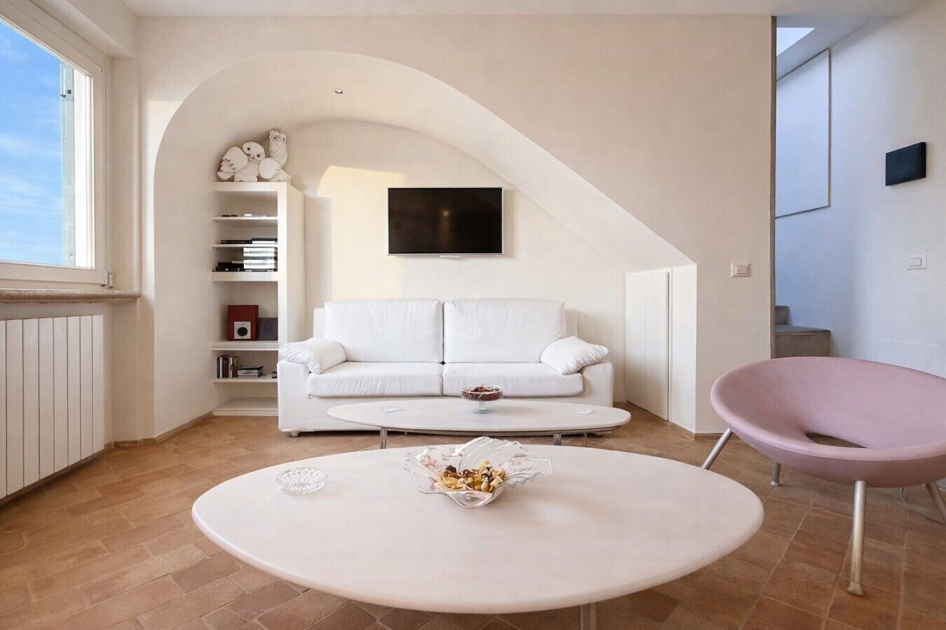 Un soggiorno moderno con un divano bianco, un tavolino rotondo, una poltrona rosa e una TV a parete.
