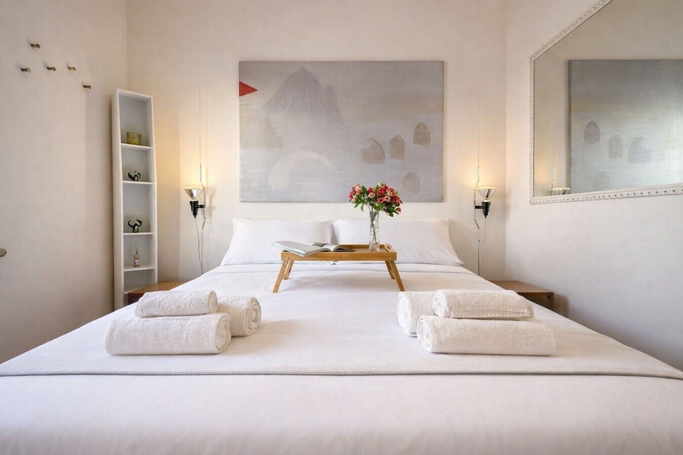 Ein Hotelzimmer mit einem Bett, einem Nachttisch mit Tablett, einer Vase mit Blumen, einem Ablagefach mit Pflegeprodukten und einem Bild an der Wand.