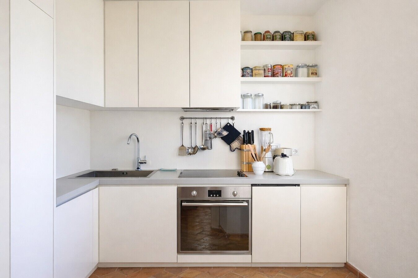 Una cucina moderna con mobili bianchi, un lavello in acciaio inossidabile, un forno integrato e una selezione di utensili ed elettrodomestici sul piano di lavoro.
