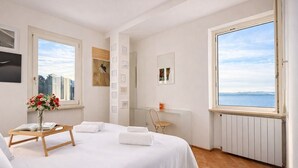 A hotel room with a bed, a small table with a vase of flowers, a chair, and a large window with a view of the sea. - Charming 2-Bedroom Home | Terrace & Great Lake View (Anguillara Sabazia)
