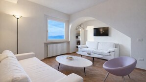 A modern living room with a white sofa, a purple chair, a white coffee table, a TV mounted on the wall, and a window with a view. - Charming 2-Bedroom Home | Terrace & Great Lake View (Anguillara Sabazia)