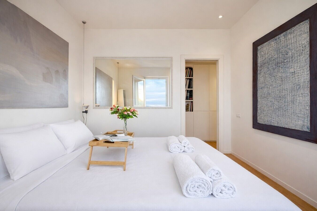 A hotel room with a bed, a bedside table with a vase of flowers, rolled towels, a large mirror, and a bookshelf.