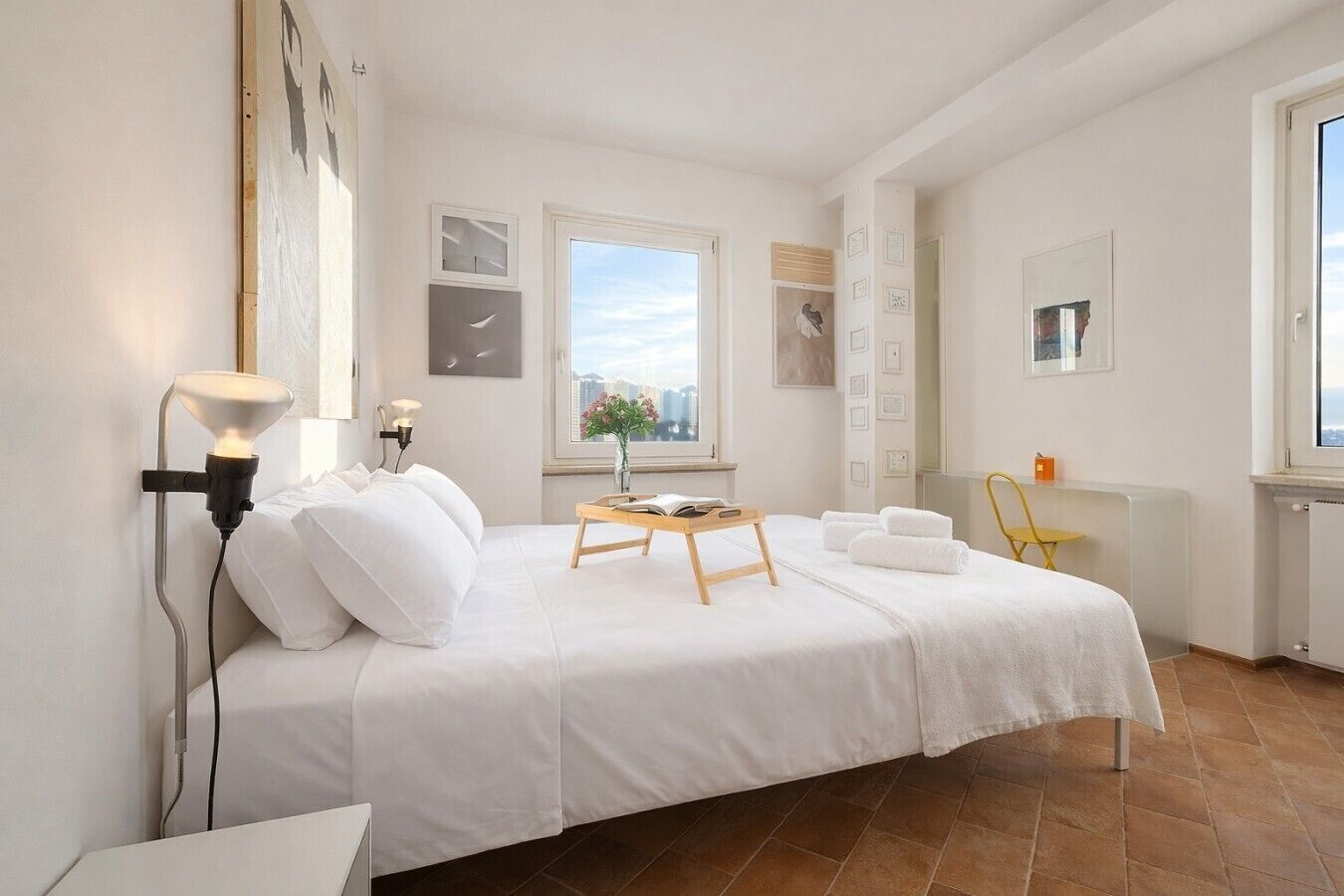 Een hotelkamer met een bed, nachtkastje, lamp, raam en kunstwerken aan de muur.