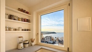 A kitchen with shelves of jars, a blender, and a window with a view of a body of water and buildings. - Charming 2-Bedroom Home | Terrace & Great Lake View (Anguillara Sabazia)