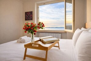 A hotel room with a bed, a bedside table with a book and a vase of flowers, and a window with a view of the sky and landscape.