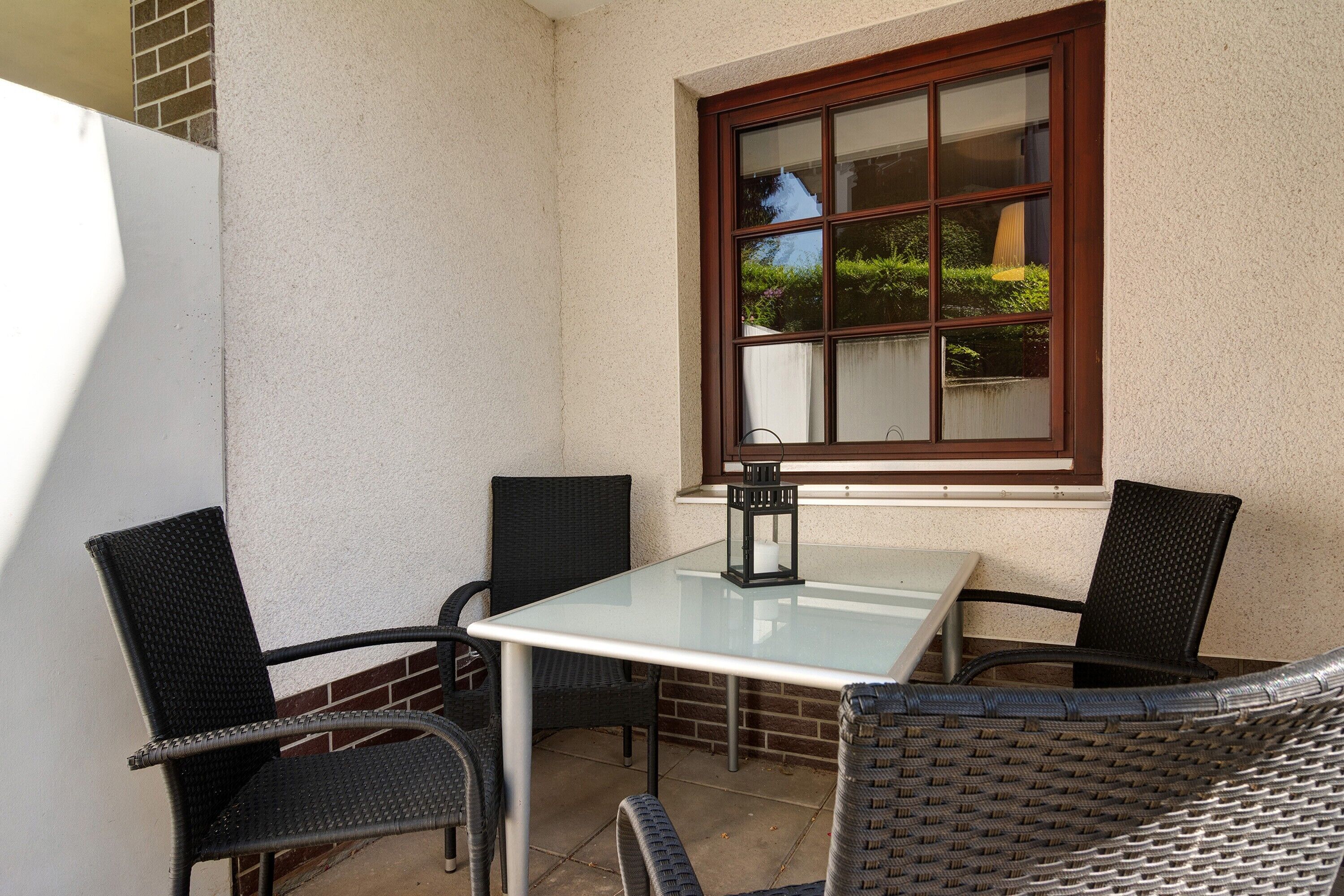 Eine Eckterrasse mit einem Glastisch und vier Korbstühlen, ein Fenster mit Blick auf Grünflächen und ein weißer Kühlschrank.