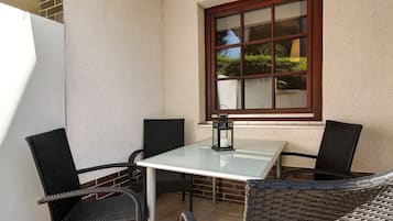 Eine Eckterrasse mit einem Glastisch und vier Korbstühlen, ein Fenster mit Blick auf Grünflächen und ein weißer Kühlschrank.