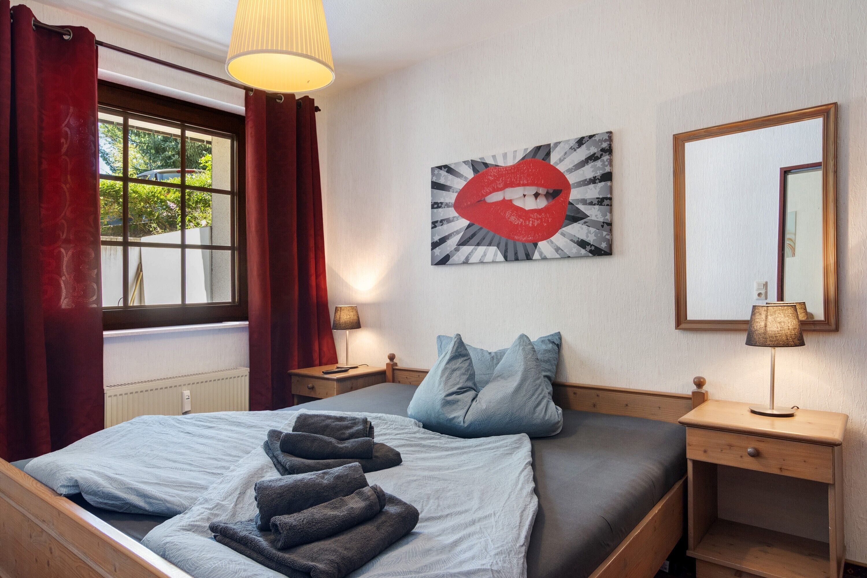 A hotel room with a bed, a nightstand, a lamp, a window with curtains, and a wall-mounted mirror.