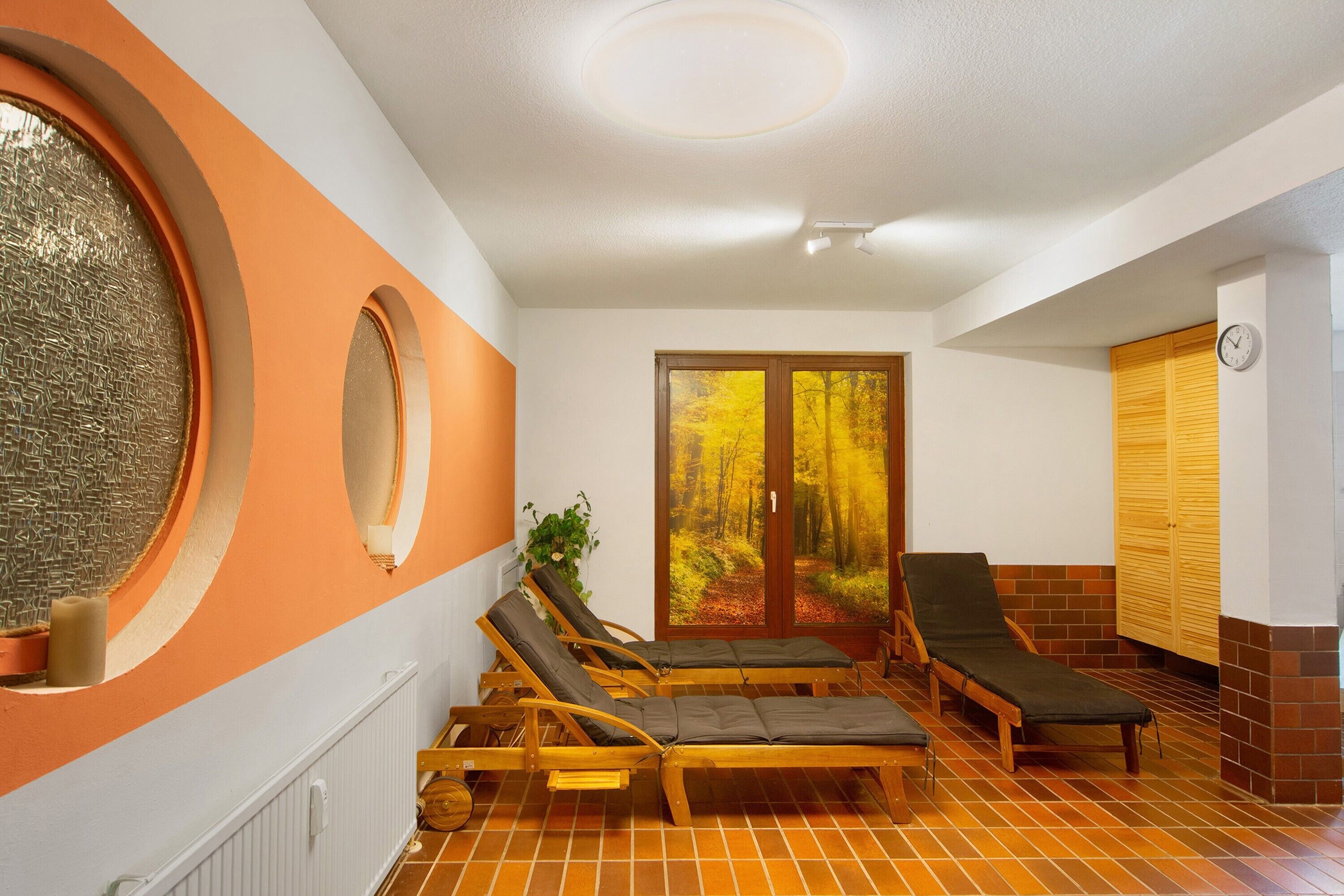 A room with two wooden lounge chairs, a window with a view of autumn trees, and a wooden door.