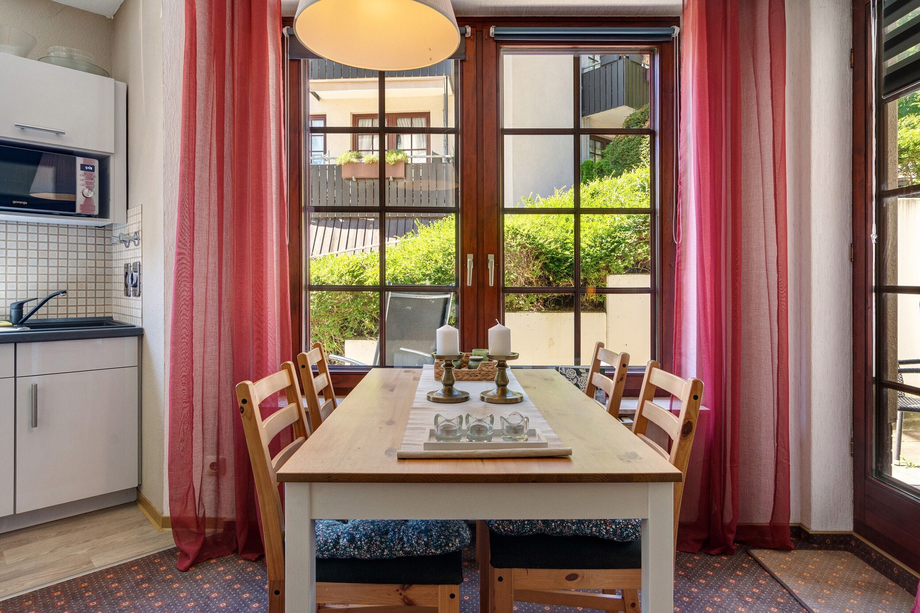 A dining area with a table set for six, a kitchen with white cabinets, and a view of greenery outside.