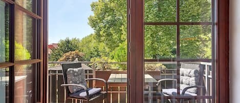 A balcony with two wicker chairs and a table, overlooking a garden with trees and a red building.